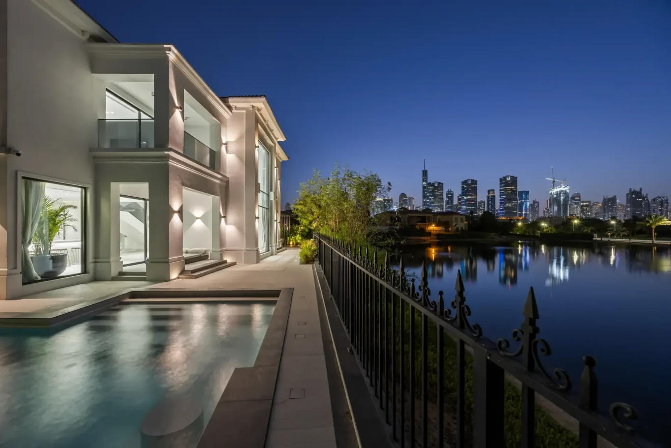 Modern white mansion with illuminated exterior, glass balconies, and a reflecting pool beside a city skyline at dusk.