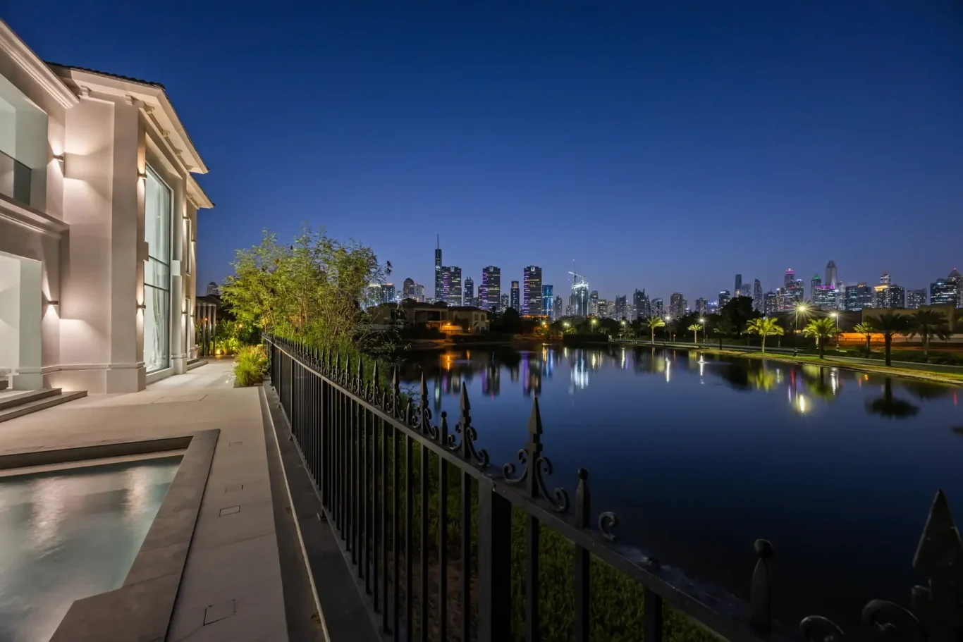 Nighttime city skyline across a calm lake, viewed from a lit modern terrace on the left with a decorative iron fence in the foreground.