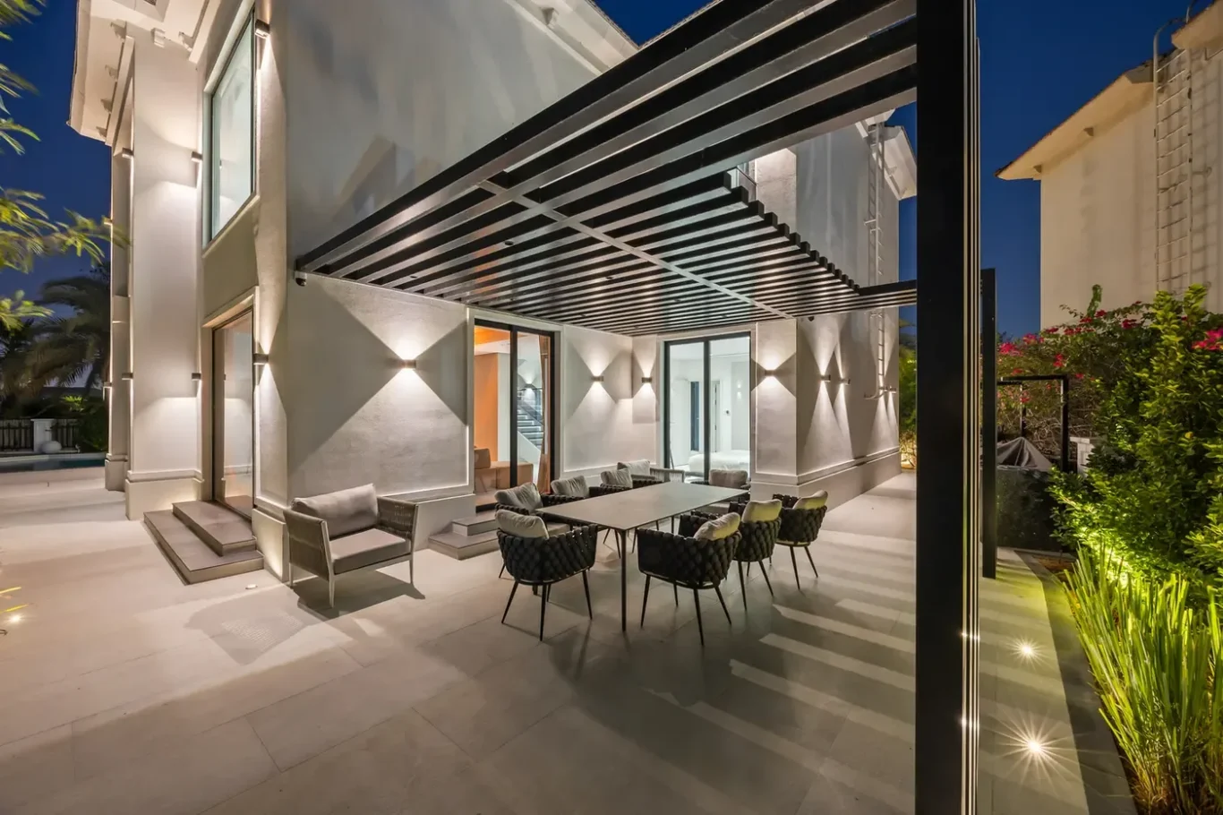 Modern back patio at night with a large outdoor dining table and woven chairs under a black slatted pergola.