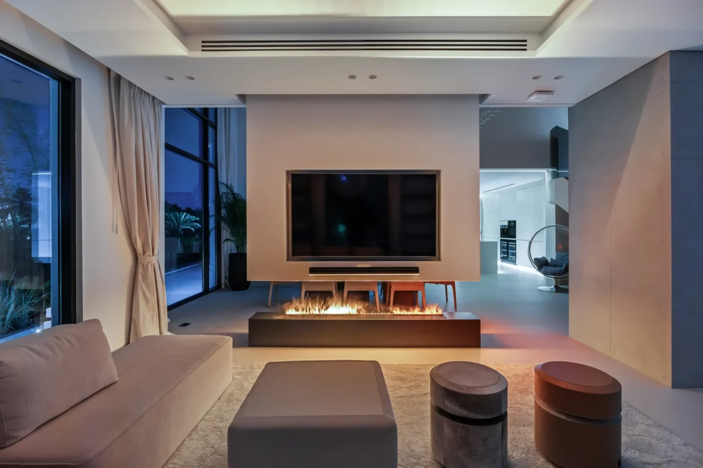 Contemporary living room with a wall-mounted TV above a linear fireplace, beige sectional sofa, and round ottomans in a spacious, modern setting at dusk.