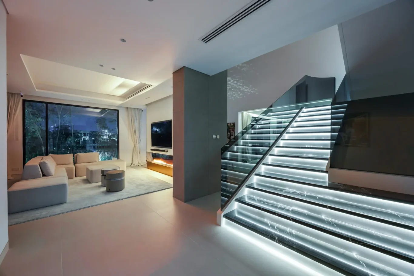 Modern living room with a white sectional sofa, floor lamp, and a glass staircase with illuminated steps beside a fireplace and large window.