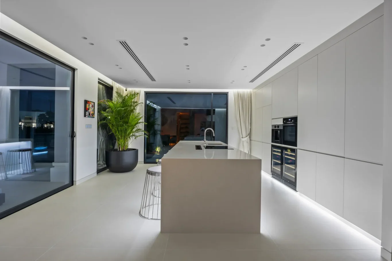 Modern white kitchen with a large central island, built-in ovens, and a sliding glass door to an outdoor area