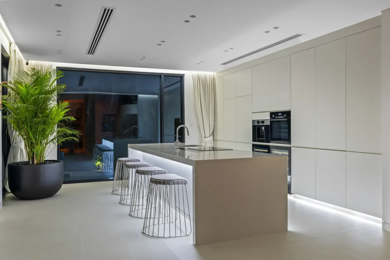 Modern white kitchen with an island, stainless steel bar stools, built-in ovens, and a tall potted plant by the glass door.