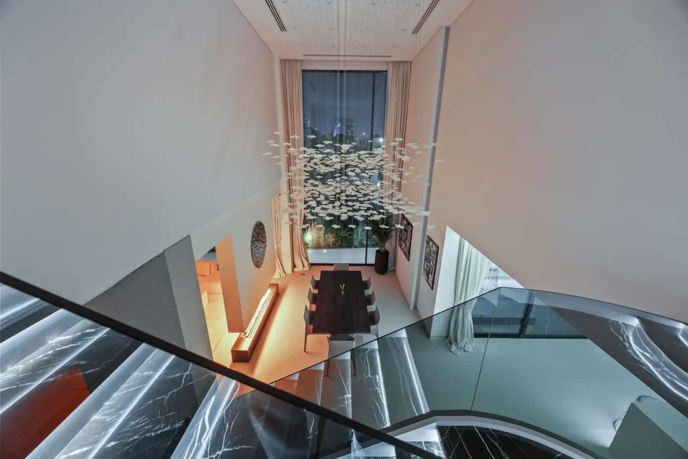 Open-concept living/dining area viewed from a glass staircase, with a long dark table and a white hanging sculpture above, plus a large window with city lights outside.
