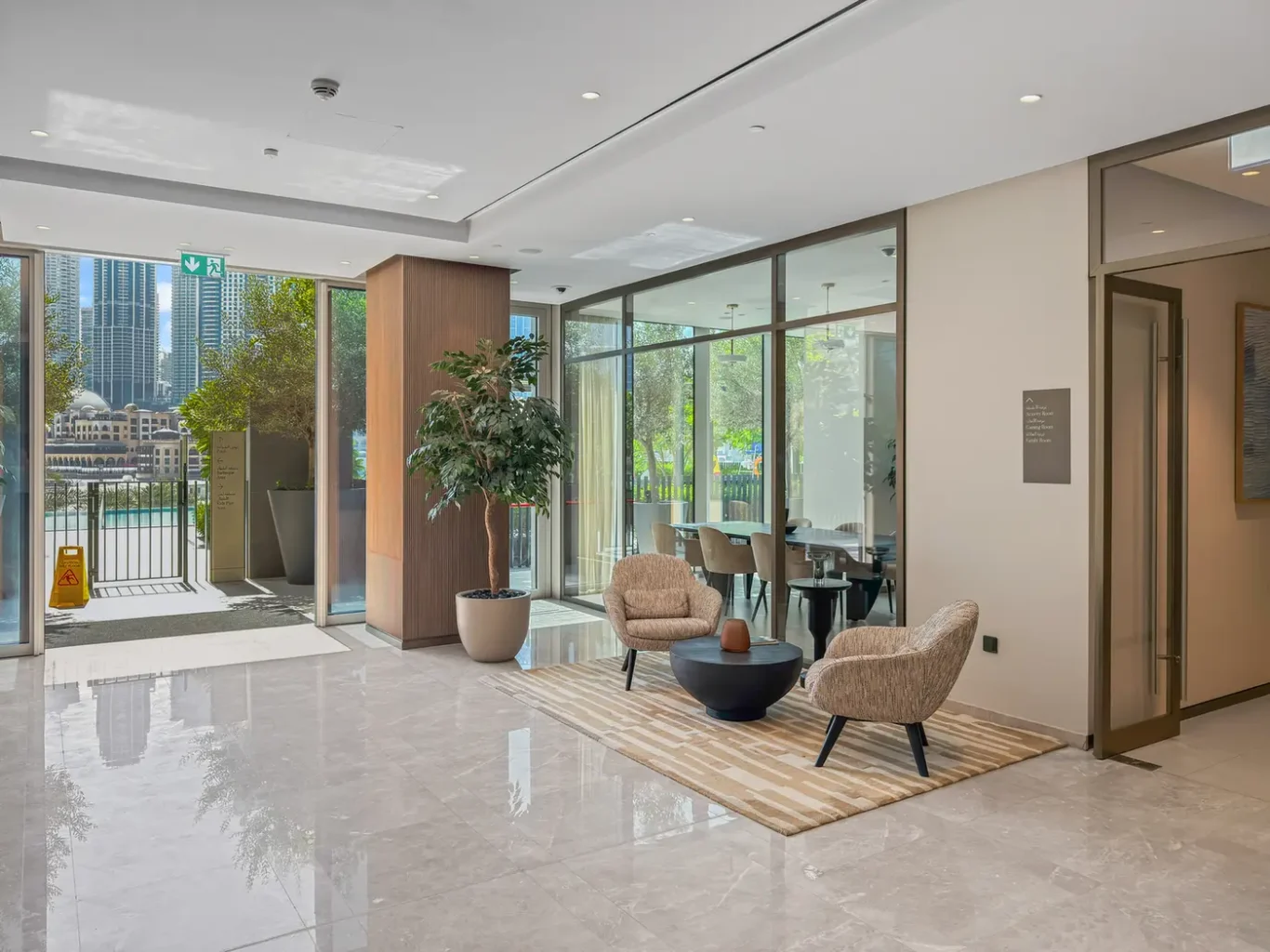 Bright hotel lobby with two beige armchairs, a round black coffee table, and a striped rug near glass walls; outdoor city view. This conveys a welcoming seating area in a modern lobby.