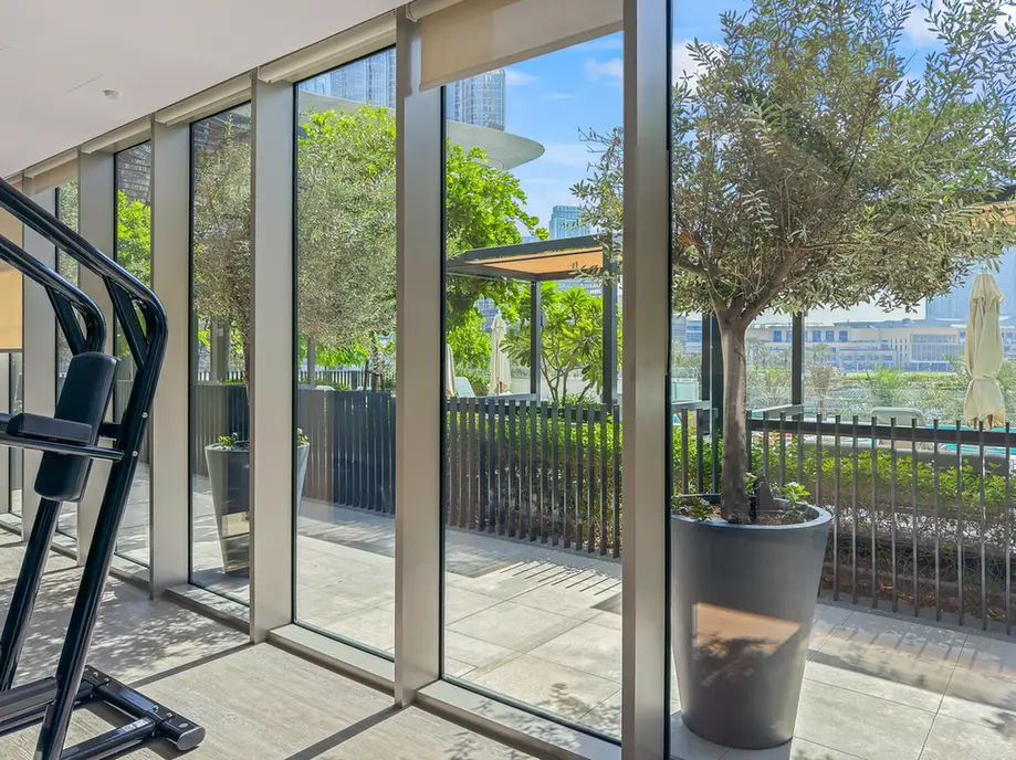 Treadmills along glass wall in a bright gym with a view of trees and city outside.