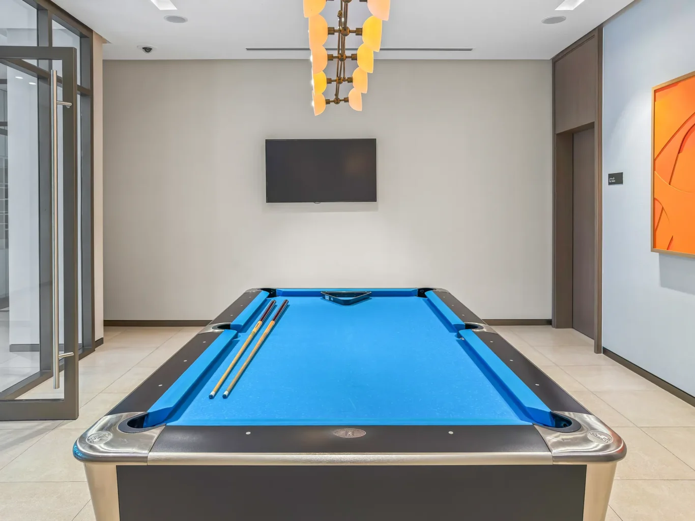 Blue billiards/pool table with two cues resting on the table, in a modern room with glass doors on the left, a wall-mounted TV on the back wall, and orange artwork on the right.