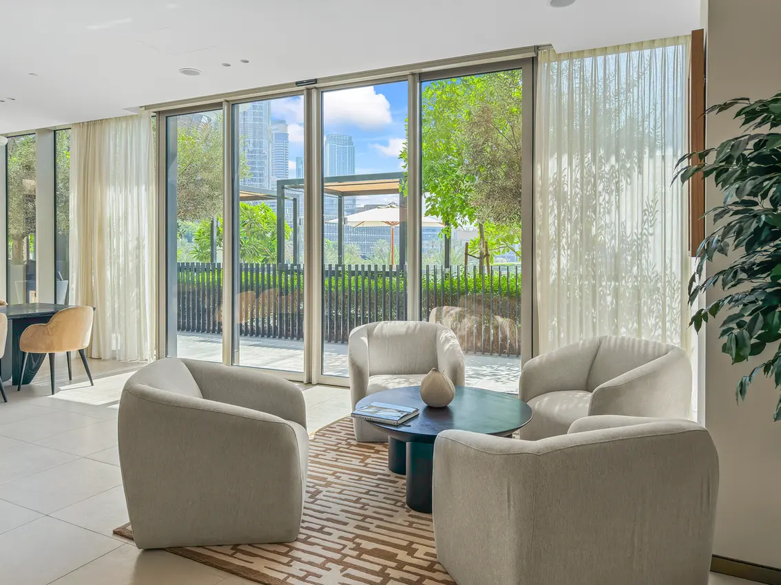 Light-filled lounge with four beige armchairs around a round blue coffee table by large glass doors opening to an outdoor patio.