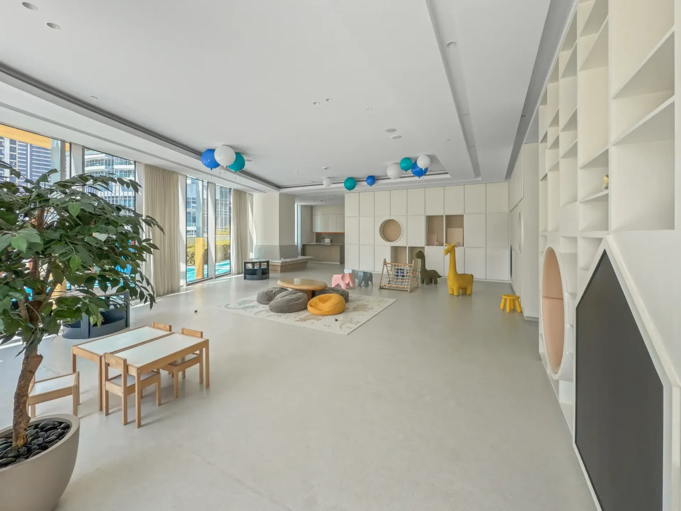 Bright modern kids' playroom with white built-in shelves, balloons, and playful animal-shaped chairs near a large windowed wall.