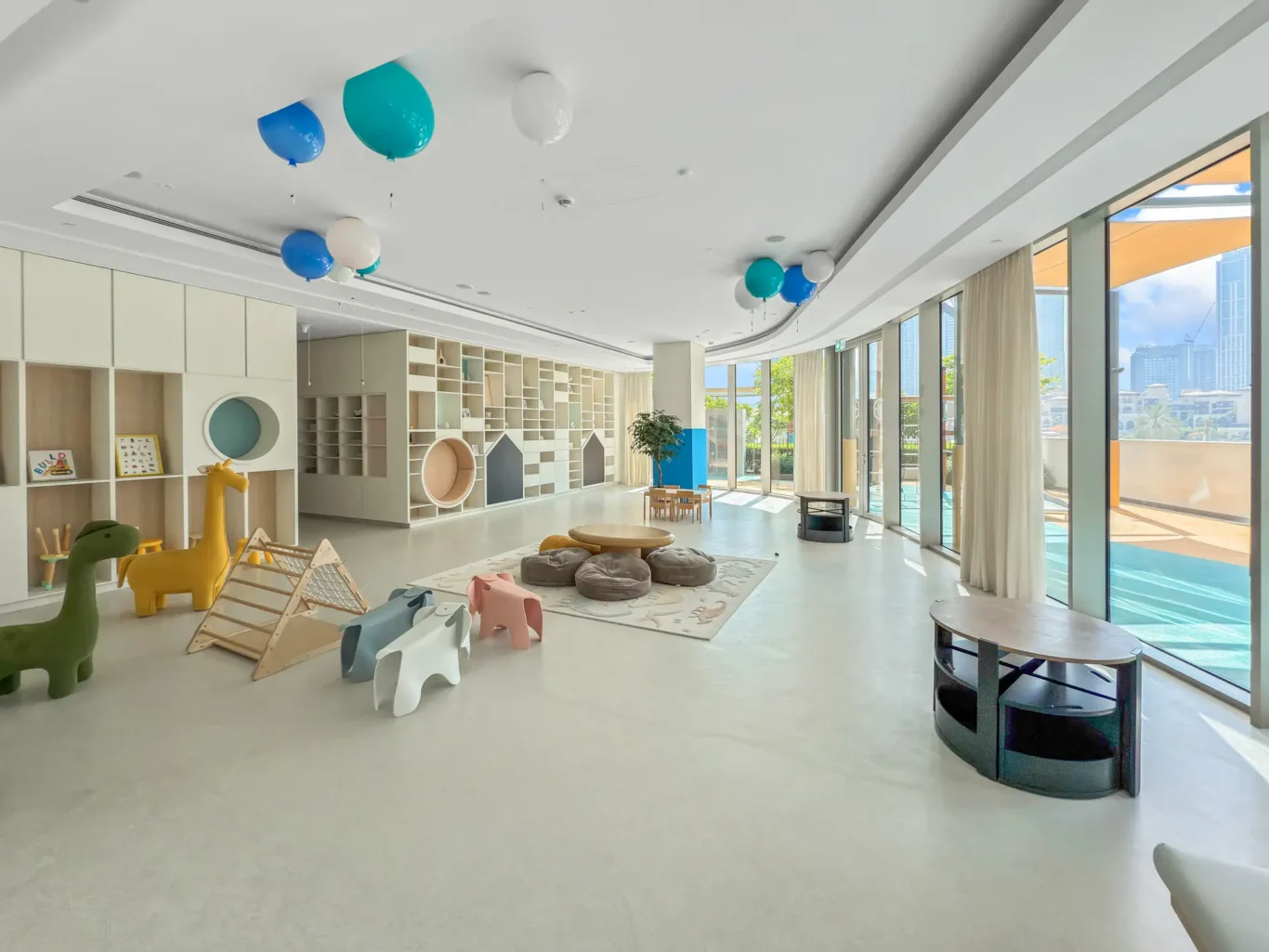 Bright, spacious kids' playroom with pastel shelving, soft seating, and colorful balloons near the ceiling; large windows with city view outside.