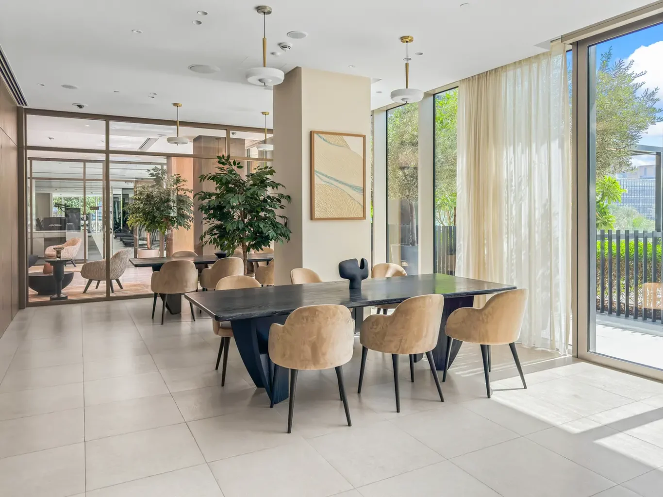 Bright modern dining area with a long dark table, beige upholstered chairs, potted plants, and floor-to-ceiling windows with sheer curtains.