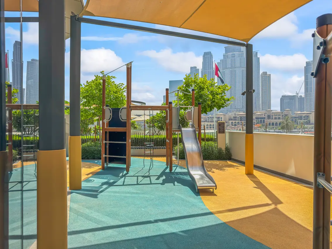 Outdoor playground on a rooftop with a metal slide and wooden climbing frame, shaded by a yellow canopy, with a city skyline in the background.