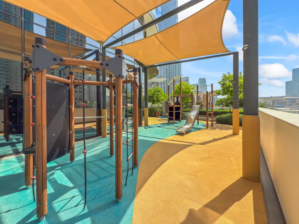 Rooftop playground with wooden climbing frames, rope elements, and a metal slide under yellow shade sails with a city skyline behind.
