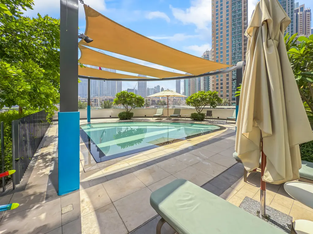Rooftop pool area with beige lounge chairs, closed umbrella, and orange shade sails against a city skyline and blue sky in the background.