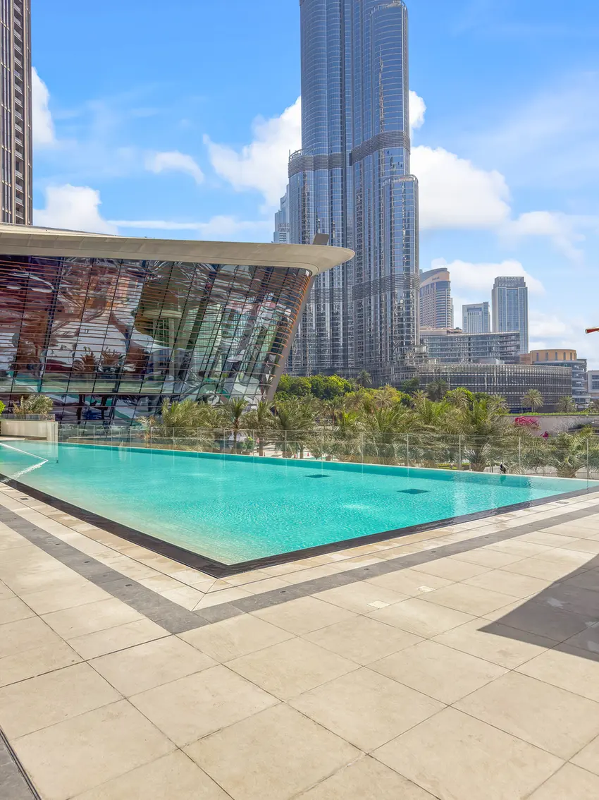 Poolside view of a turquoise swimming pool with a glass-fronted, angled building and towering skyline in a sunny urban area