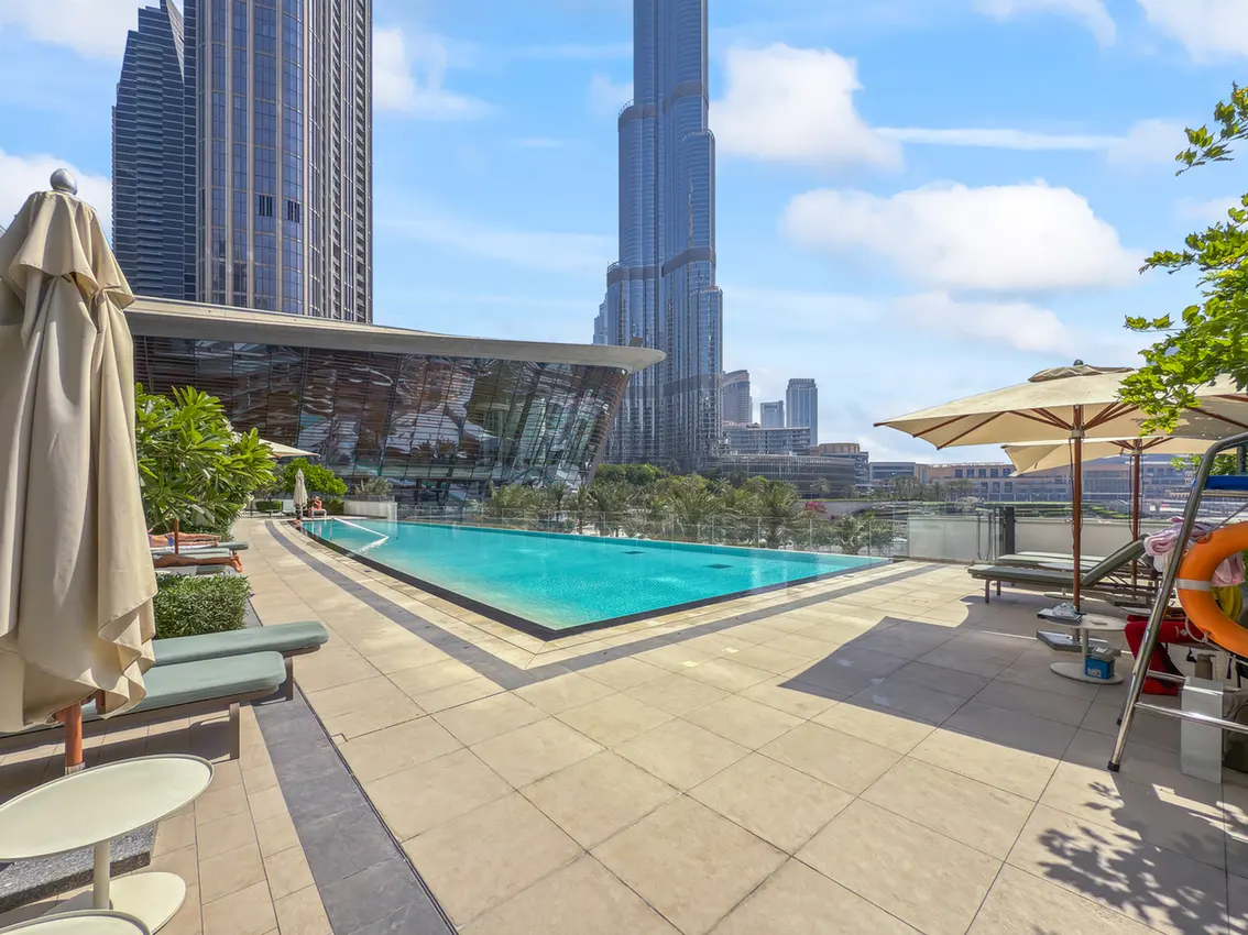 Rooftop pool deck with lounge chairs and beige umbrellas, overlooking a city skyline of tall glass skyscrapers under a blue sky.
