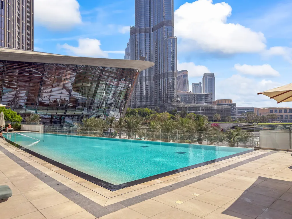 Rooftop swimming pool with turquoise water and glass railings, a curved glass building nearby, and a skyline of tall buildings under a bright sky.