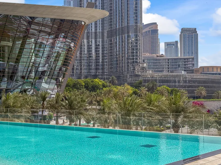 Modern city skyline with glass skyscrapers and a curved architectural pavilion overlooking a turquoise pool and palm trees.