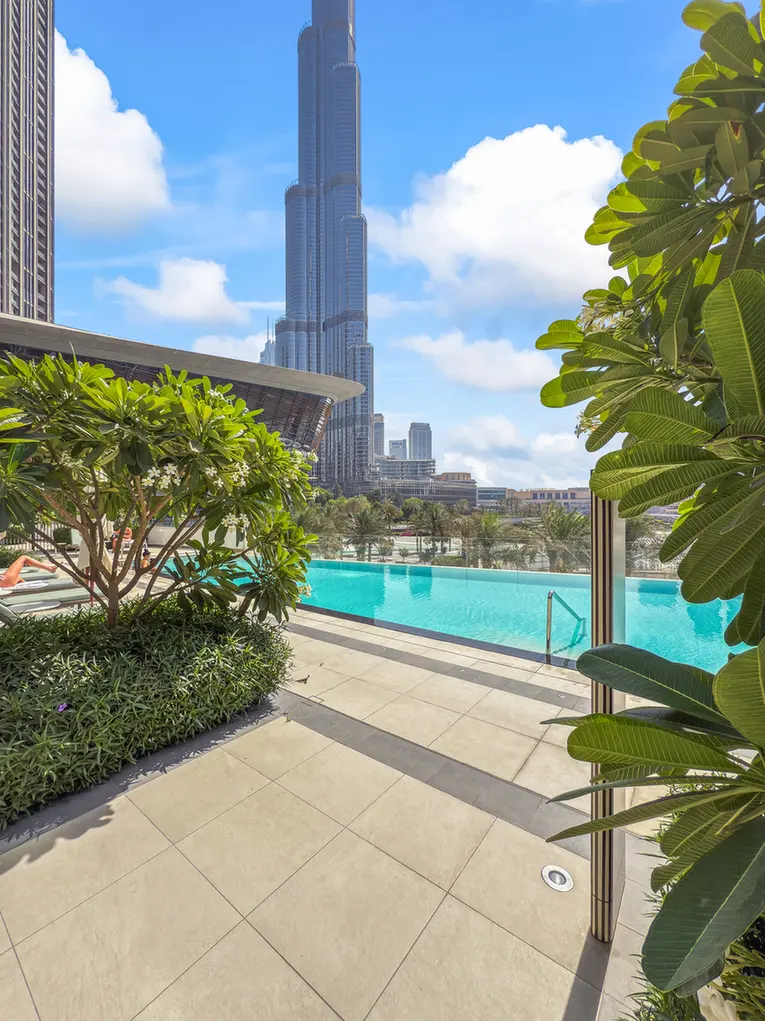 Poolside terrace with a bright blue swimming pool and lush plants, skyscrapers visible in the background under a sunny sky.