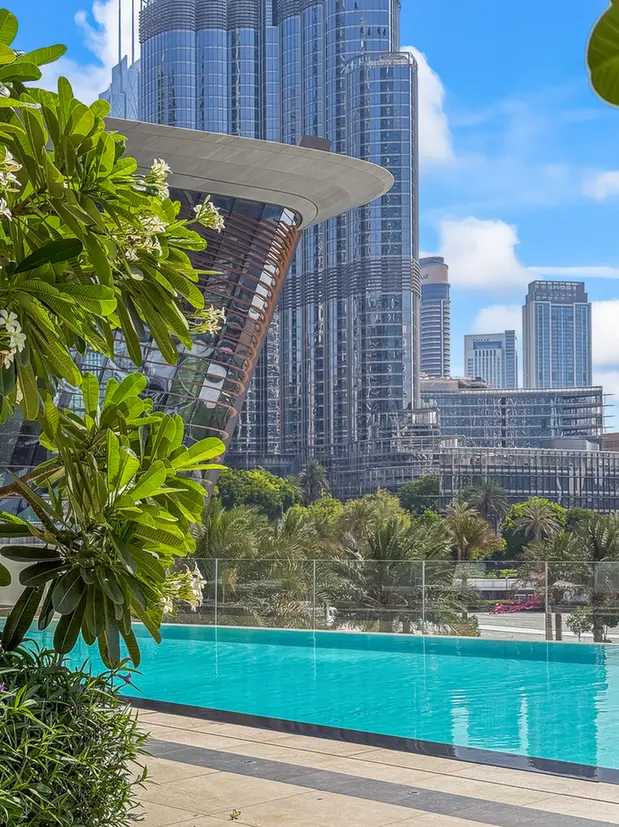 Hotel rooftop pool with turquoise water and glass railing, city skyscrapers in the background under a blue sky with greenery in the foreground.
