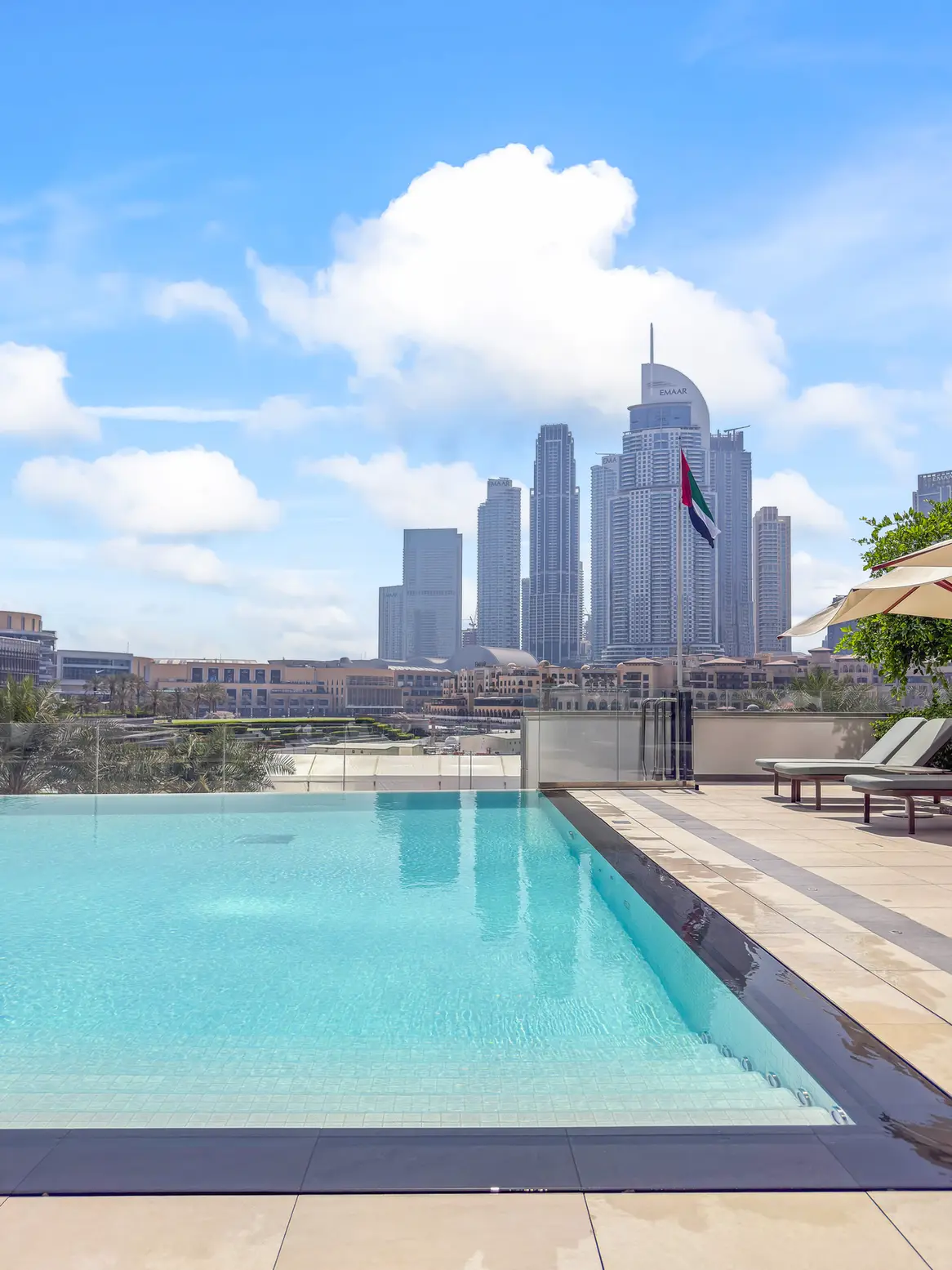 Poolside on a sunny terrace with turquoise water and loungers, overlooking a cluster of tall UAE skyscrapers with a visible UAE flag in the foreground.