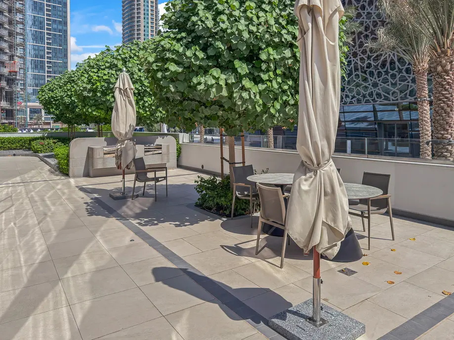 Outdoor coworking plaza with round tables and beige umbrellas, shaded by trimmed trees and urban high-r rises in the background.