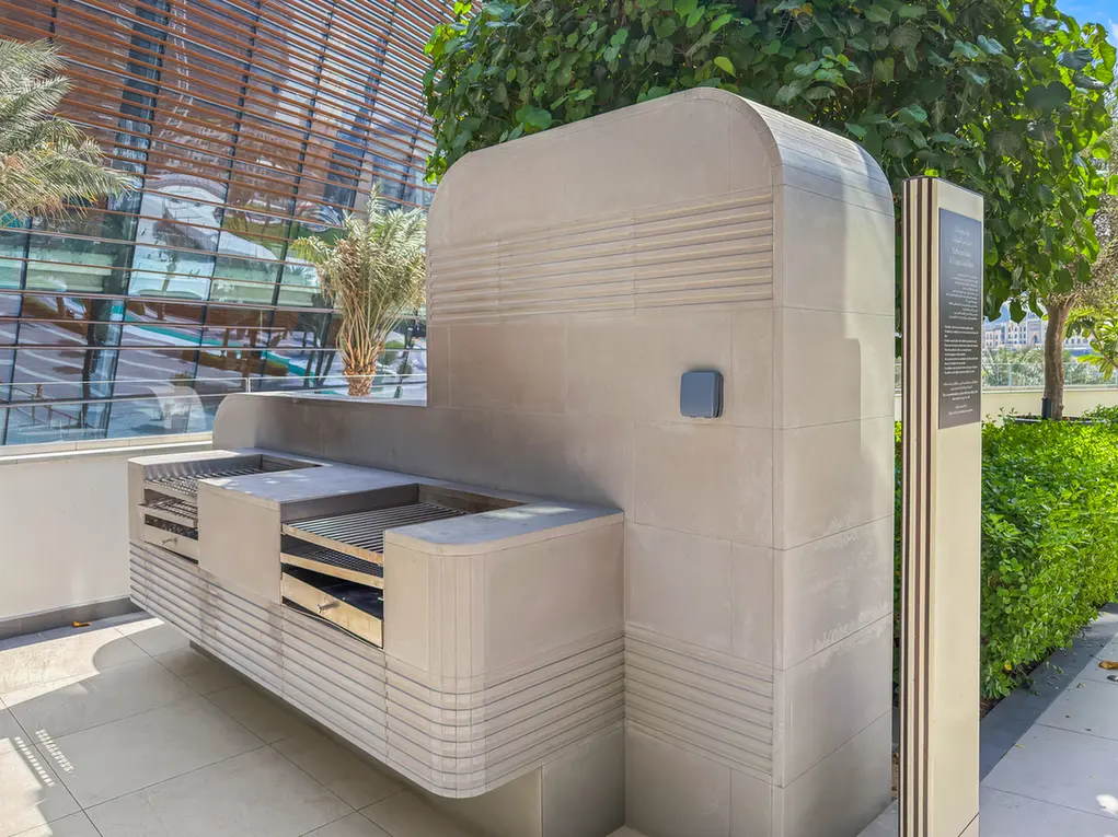 Built-in stainless steel outdoor grill station with shelving, set in a sunny courtyard against a glass-walled building and green plants.