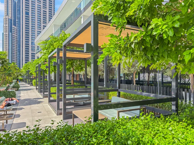 Outdoor seating pods with beige canopies along a sunlit courtyard, surrounded by greenery and city skyscrapers nearby.
