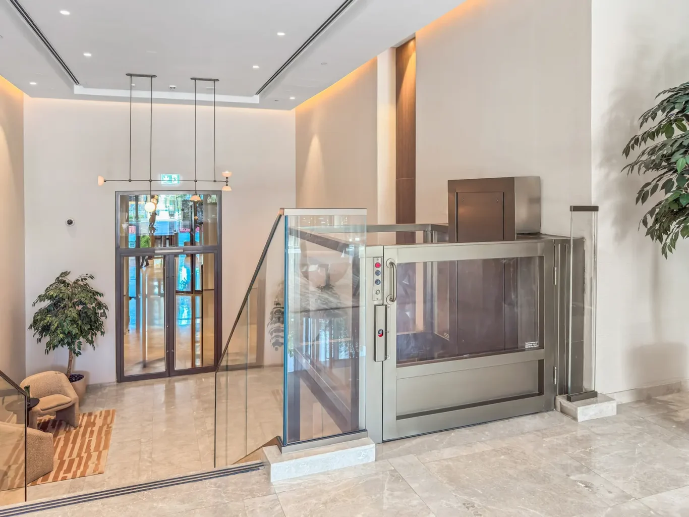 Modern lobby with a glass-encased elevator, glass railing, and potted plants.
