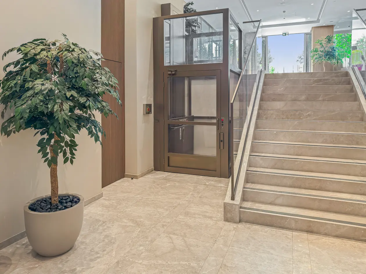 Indoor lobby with a brown-framed glass elevator near a staircase and a large potted plant on marble flooring