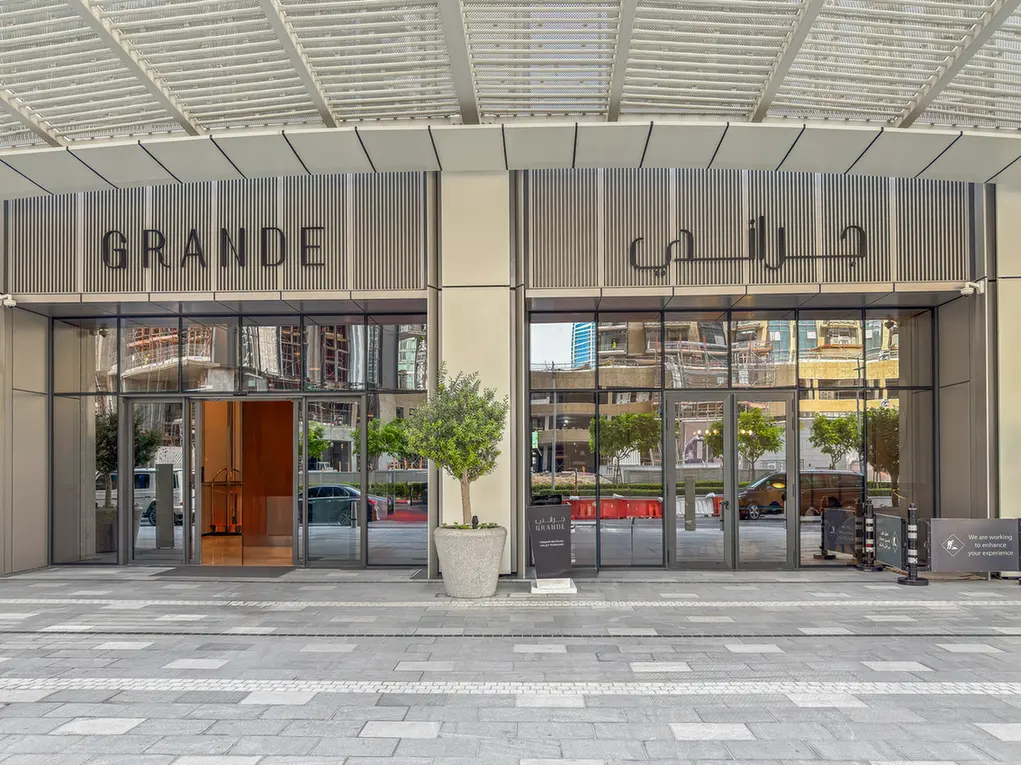 Front entrance of a modern building named 'GRANDE' with glass doors, a potted tree, and Arabic signage, reflecting a city street in the glass panes.