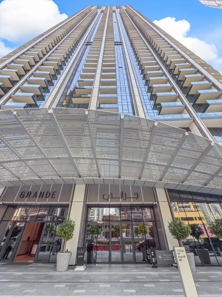 Low-angle view of a tall modern high-rise with glass and balconies, entrance under a metal canopy labeled 'GRANDE'.
