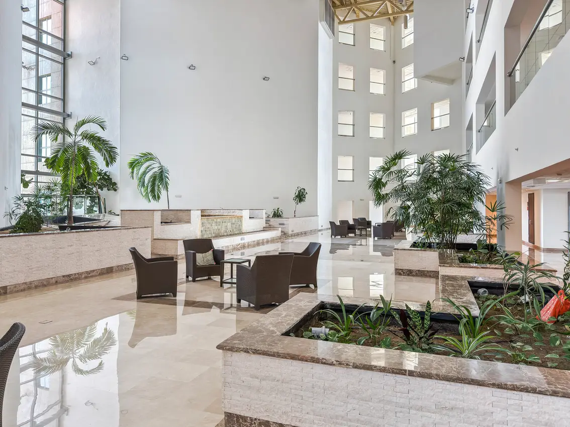 Bright hotel lobby with tall windows, white walls, and brown seating around a water feature.