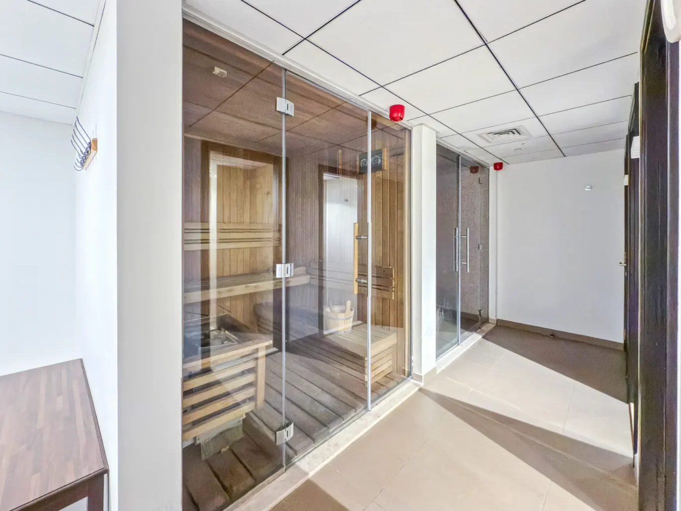 Glass-enclosed sauna with wooden benches in a hallway, red emergency lights above the room, tiled floor nearby.