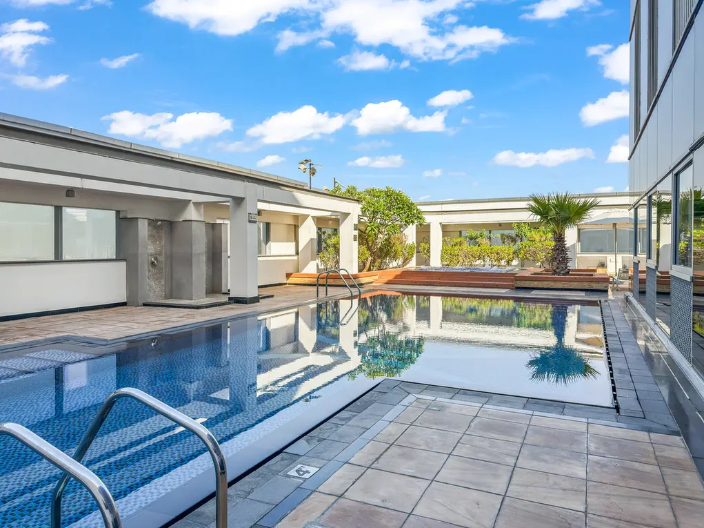Rooftop pool area with blue water, lounge benches, a palm tree, and modern glass-and-stone buildings under a sunny sky.