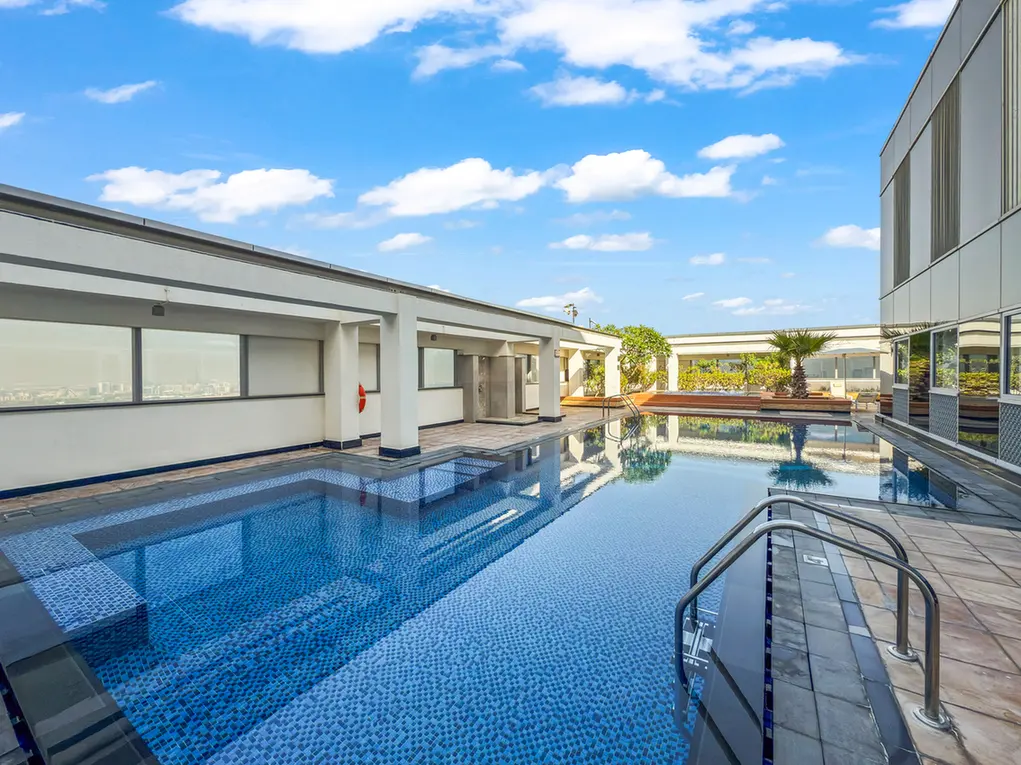 Rooftop swimming pool on a modern building with blue-tiled water and clear sky, glass railing on the right, and lounge areas in the background.