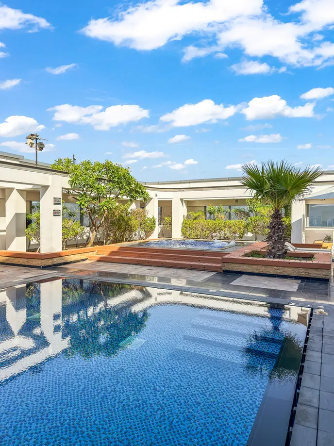 Rooftop pool area on a sunny day with palm trees and modern white buildings surrounding blue water. A clear sky with scattered clouds above.