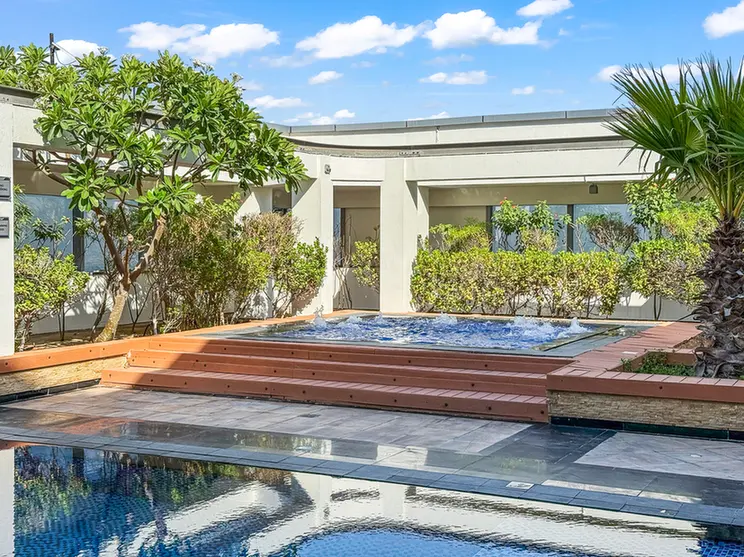Hotel courtyard with a rectangular pool, wooden steps, and lush green shrubs under a blue sky.