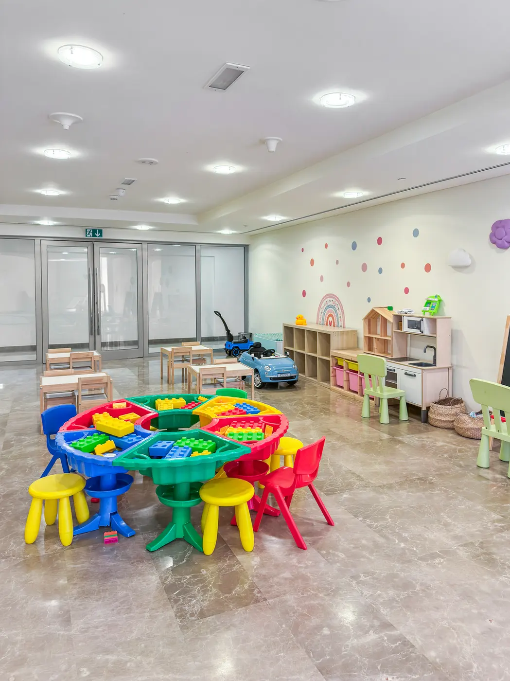 Colorful children's playroom with a round multi-colored table, small stools, and toy blocks; shelves and a pretend kitchen along the wall.