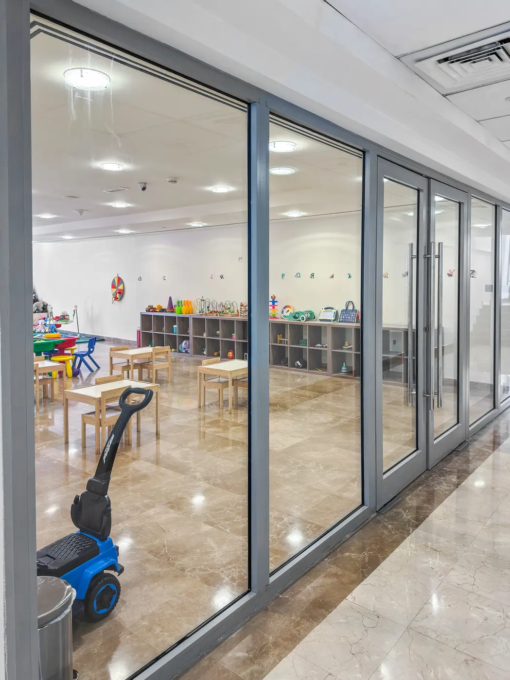 Playroom for children seen through glass storefront with small tables, shelves of toys, and a blue ride-on toy in the foreground.
