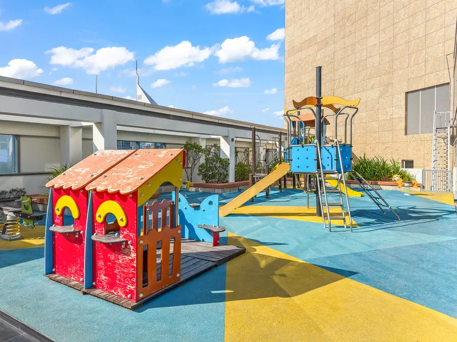 Colorful rooftop playground with a red wooden playhouse, yellow slide, and blue climbing structures on blue-yellow safety flooring.
