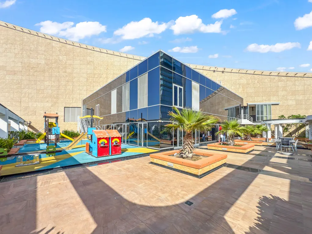 Outdoor modern facility with glass-walled building, wooden planters, palm trees, and a colorful children's playground on a sunny day.