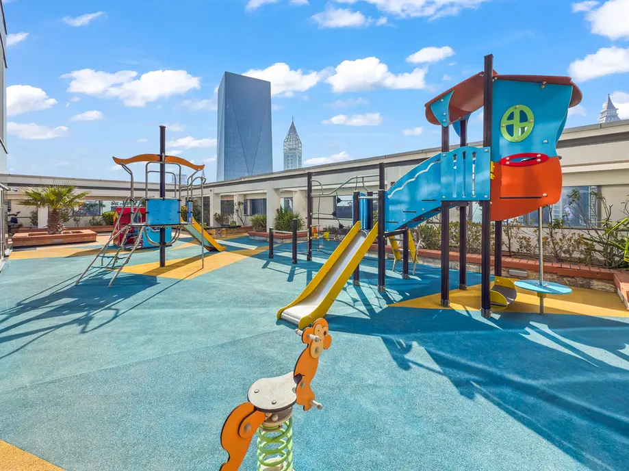 Colorful rooftop playground with slides and climbing structures on a bright day, skyline of tall buildings in the background.