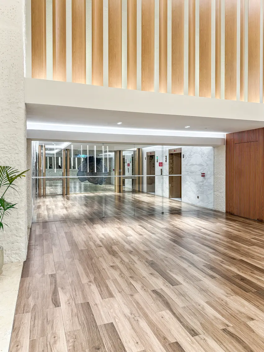 Modern office lobby with a wooden slat ceiling, glass doors, marble walls, and an elevator bank visible ahead.