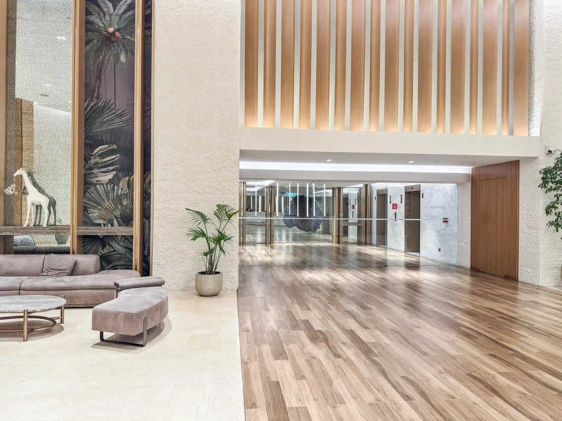 Contemporary hotel lobby with beige seating, a round coffee table, and a potted plant near a palm mural panel.