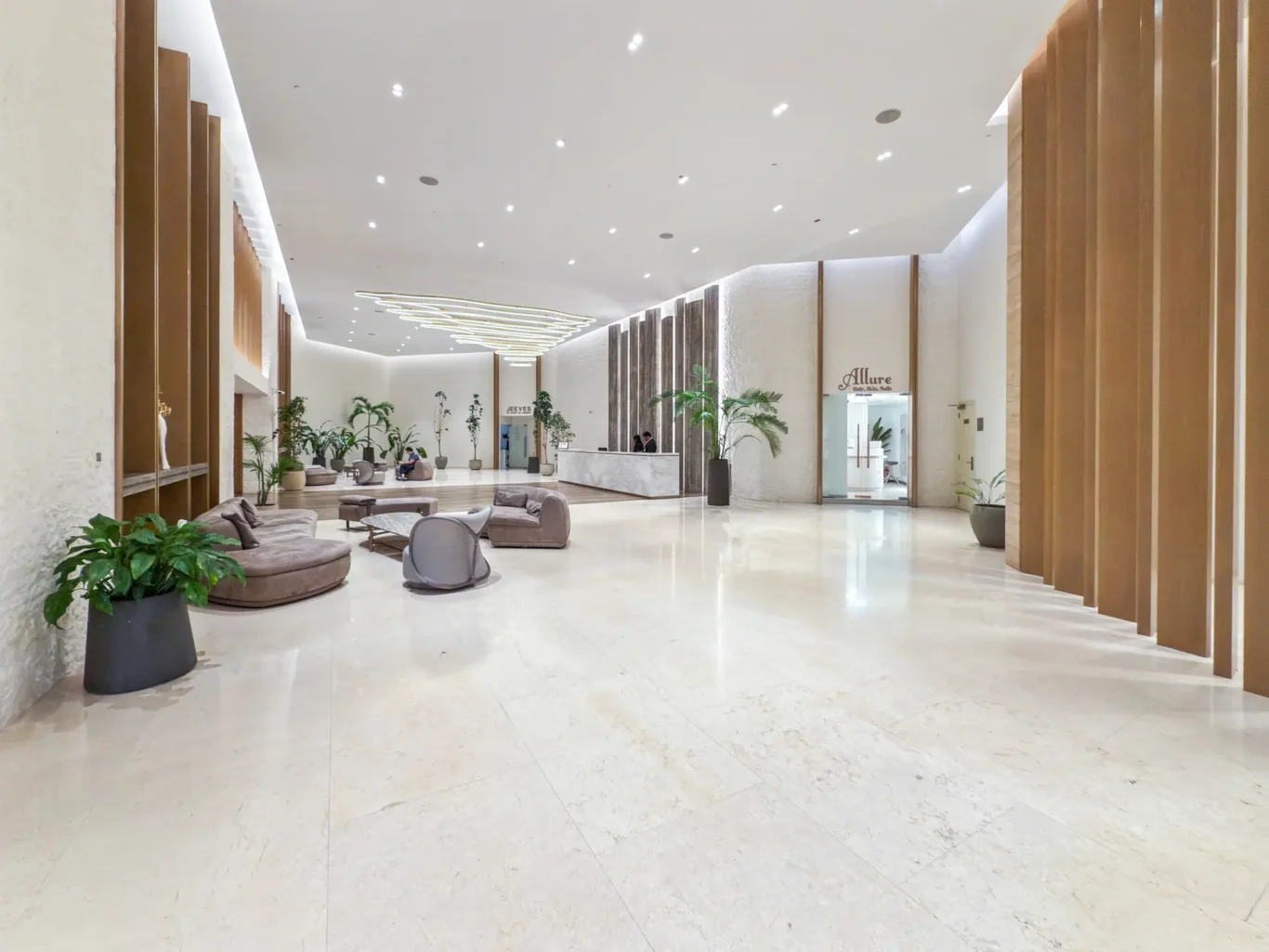 Spacious modern hotel lobby with marble floors, circular brown seating, and potted plants lining a wide hall.