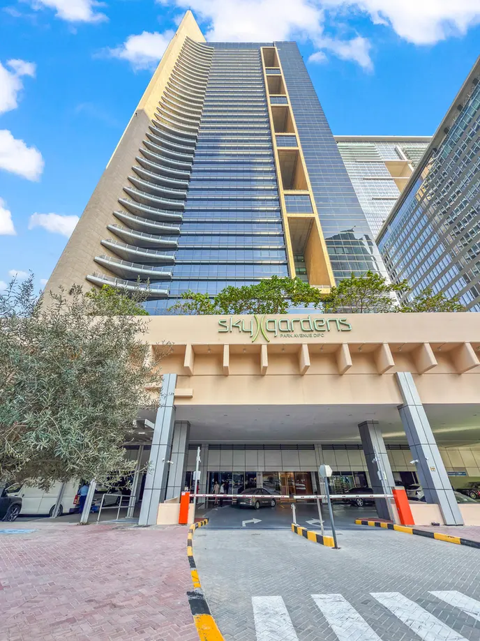 Tall glass high-rise with curved balconies and a beige entrance area labeled 'Sky Gardens' at the base, driveway and pedestrians visible beneath.