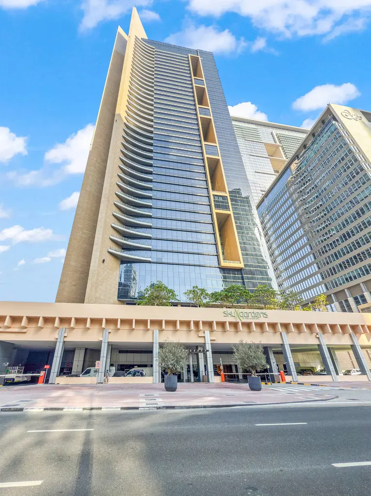 Tall glass high-rise with curved balconies and beige accents, front plaza and street below under a blue sky.