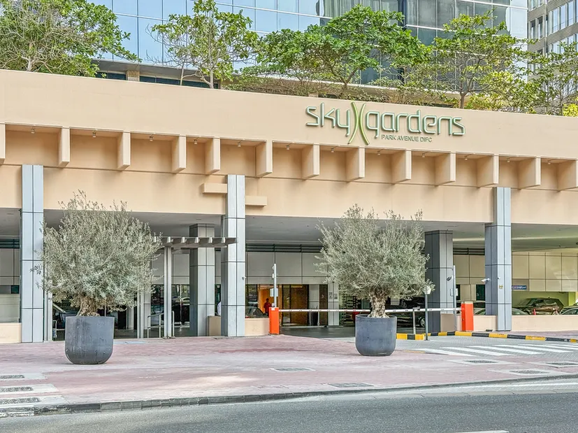 Entrance to SkyGardens building with two large potted trees, glass doors, and a driveway barrier.