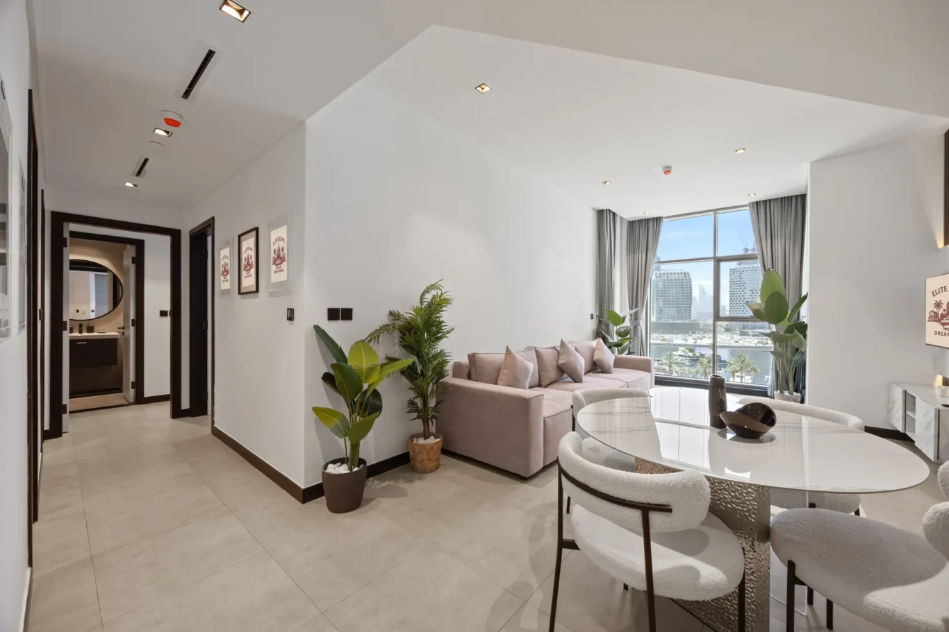 Spacious living room with a beige sectional, glass dining table, and potted plants by large windows overlooking the city skyline.